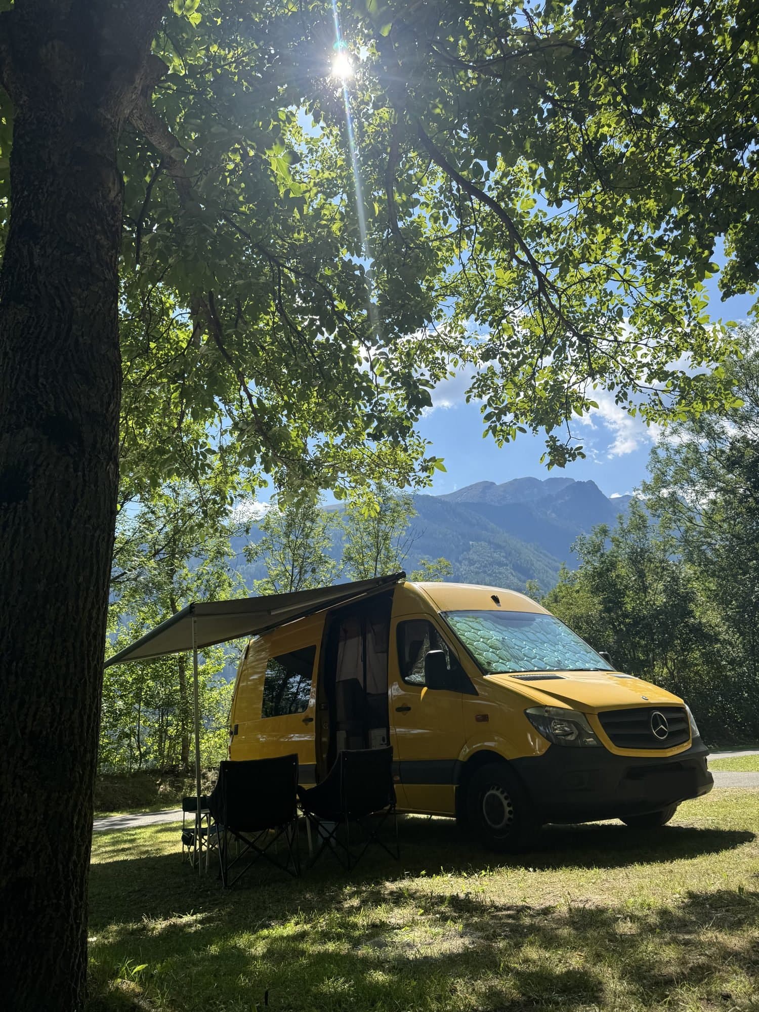 Žluťák, a yellow Mercedes-Benz Sprinter camper with its awning out, parked under a tree with mountains behind